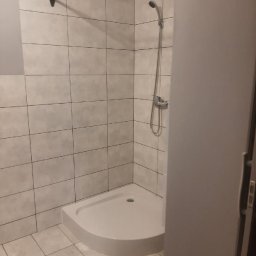 Ryszard Łaszewski - Narożny brodzik z kabiną prysznicową wyłożoną jasnymi, kwadratowymi płytkami z ciemniejszą fugą, widok zza częściowo otwartych drzwi.