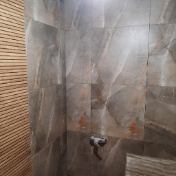 Ryszard Łaszewski - Fragment łazienki z ułożonymi płytkami imitującymi marmur, widoczny surowy zawór i fragment ściany wykończonej drewnianymi listwami.