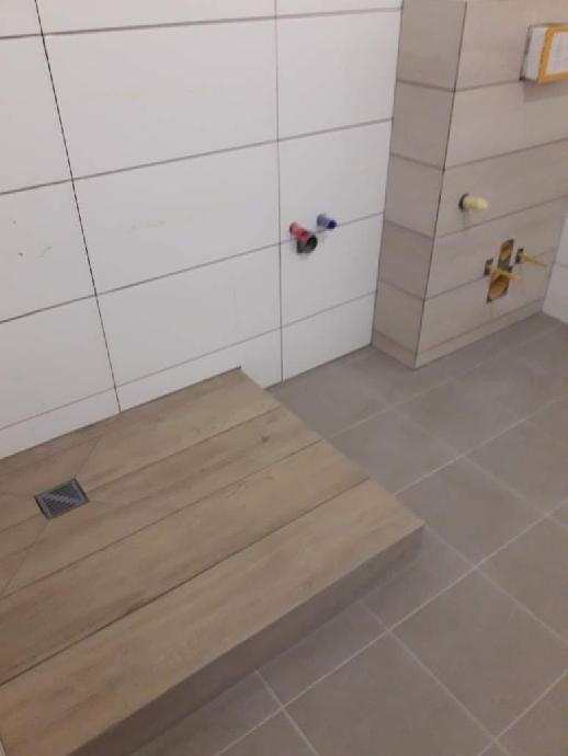 Płytki ceramiczne imitujące drewno na podłodze prysznica z odpływem liniowym, białe płytki ścienne z wyprowadzonymi instalacjami wodnymi.