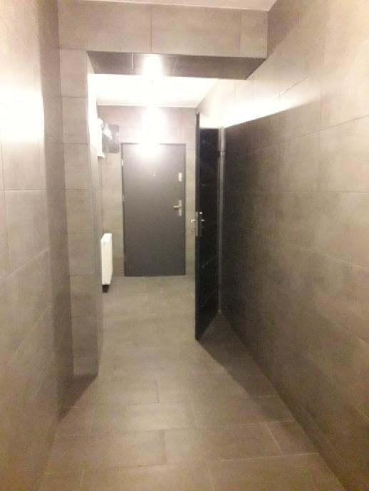 Korytarz wykończony szarą płytką ceramiczną na ścianach i podłodze, z widocznym fragmentem otwartych, ciemnych drzwi po prawej stronie i zamkniętymi szarymi drzwiami na wprost.