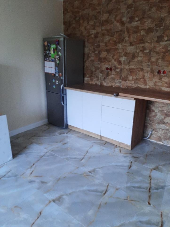 Kuchnia w trakcie wykańczania z marmurową podłogą, białe szafki z drewnianym blatem i ściana wyłożona kamieniem dekoracyjnym, obok stoi srebrna lodówka.