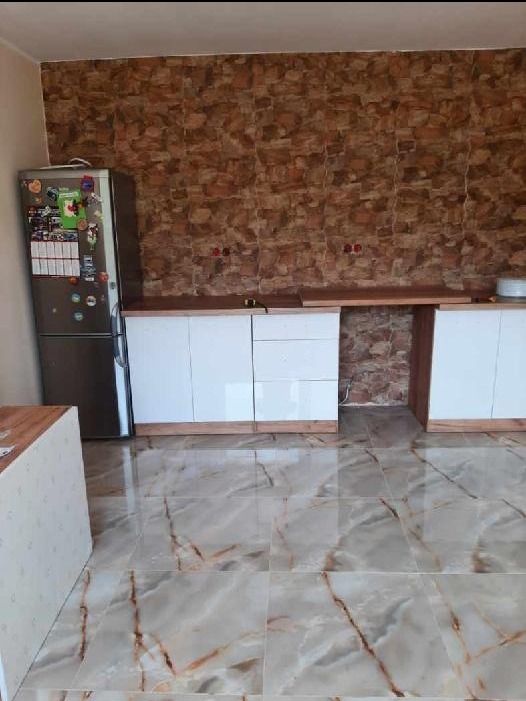 Kuchnia z białymi szafkami, blatem z drewna, ścianą wyłożoną kamieniem dekoracyjnym i marmurową podłogą, w tle lodówka z magnesami.