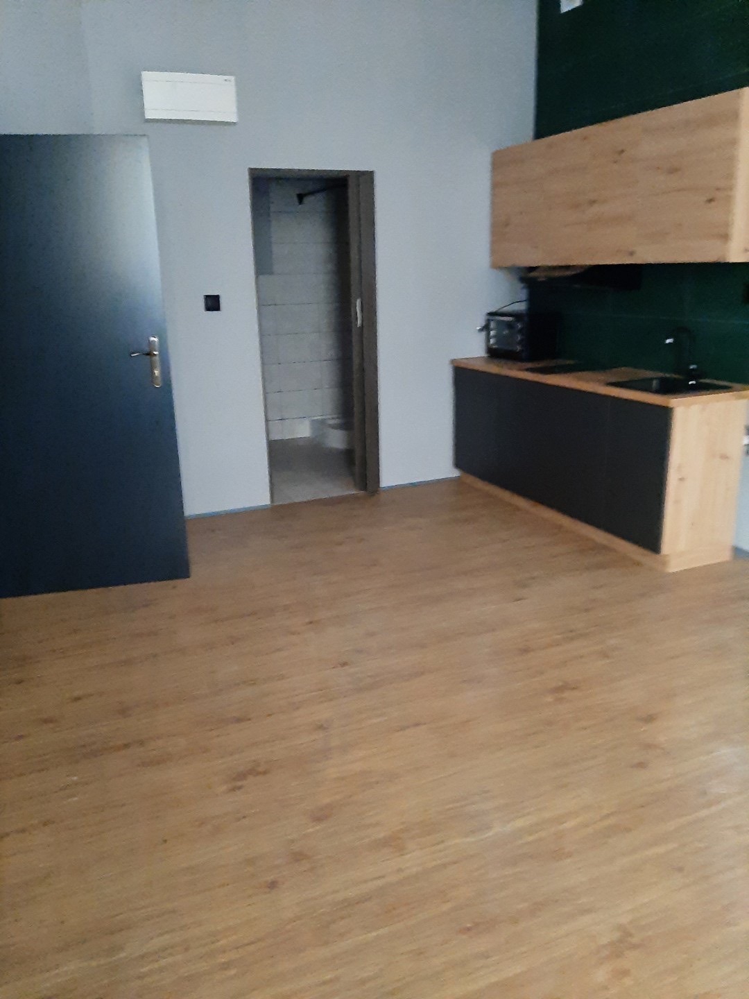 Wnętrze po remoncie: widok na nową podłogę z paneli, drzwi wejściowe, oraz zabudowę kuchenną z czarnymi frontami i drewnianymi szafkami wiszącymi.