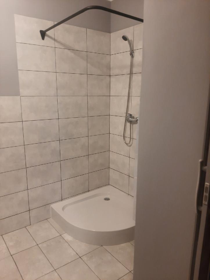 Narożny brodzik z kabiną prysznicową wyłożoną jasnymi, kwadratowymi płytkami z ciemniejszą fugą, widok zza częściowo otwartych drzwi.