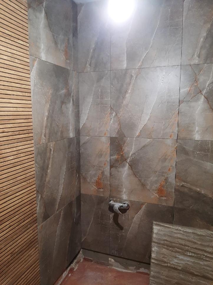 Fragment łazienki z ułożonymi płytkami imitującymi marmur, widoczny surowy zawór i fragment ściany wykończonej drewnianymi listwami.