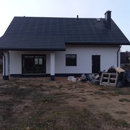 Gerbud - Biały dom jednorodzinny z ciemną dachówką, widok z zewnątrz, teren wokół domu w trakcie zagospodarowania, widoczne materiały budowlane i odpady.