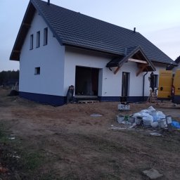 Gerbud - Biały dom jednorodzinny z ciemną dachówką w trakcie prac elewacyjnych, widoczne wiadra z farbą i pracownik na drabinie przy tylnej ścianie budynku.