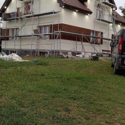 Gerbud - Dom z rusztowaniami podczas prac elewacyjnych. Część ściany z brązowymi panelami, reszta w jasnym kolorze. Widoczne worki i narzędzia.