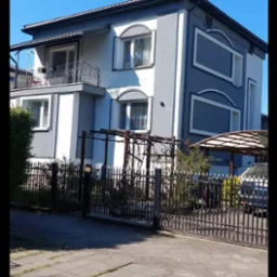 Dwukondygnacyjny dom z szaro-białą elewacją, balkonem z kutą balustradą i dekoracyjnymi obramowaniami okien. Widok z zewnątrz, brama i podjazd.