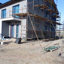 Gerbud - Dom w trakcie ocieplania elewacji. Widoczne rusztowania, materiały izolacyjne i okna zabezpieczone folią. Na ziemi narzędzia i odpady budowlane.