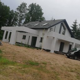 Gerbud - Nowoczesny dom jednorodzinny z białą elewacją, czarną dachówką i dużymi oknami, widziany z poziomu gruntu na tle drzew i pochmurnego nieba.