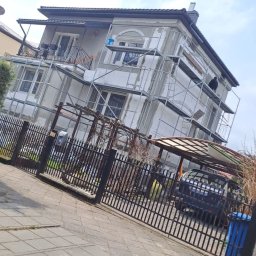 Gerbud - Dom w trakcie renowacji elewacji, z rusztowaniami i pracownikiem na górze. Widoczne ogrodzenie, chodnik i samochód w tle.