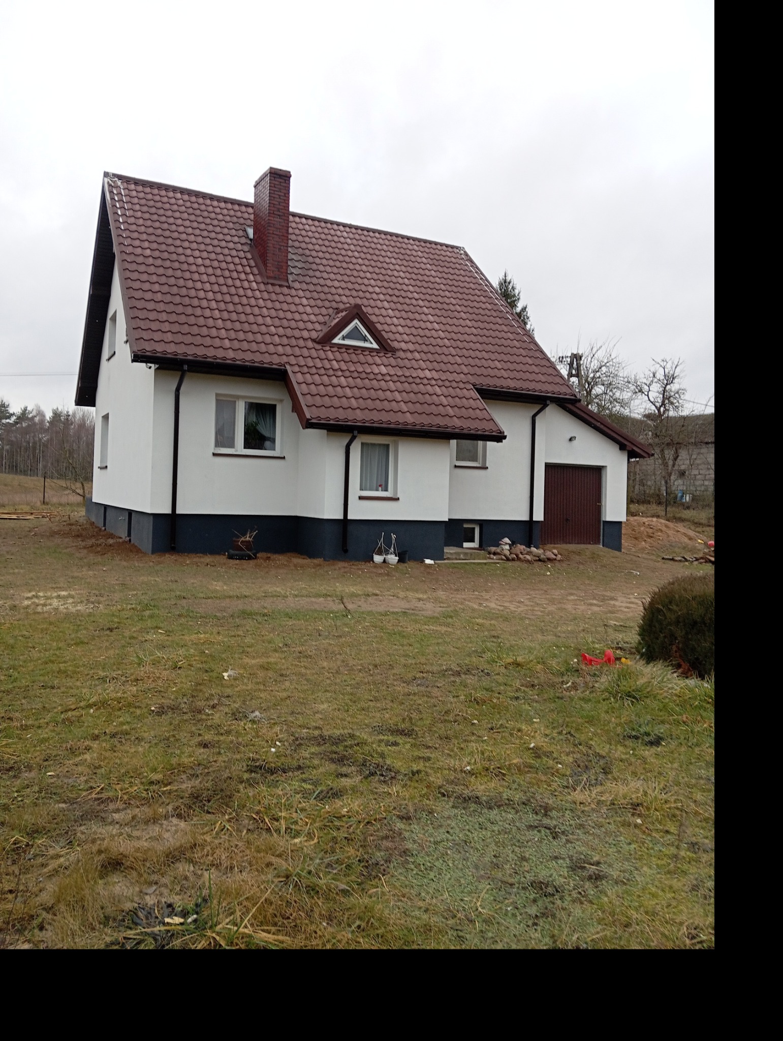 Dom z białą elewacją i brązowym dachem, widok z boku. Ciemna podmurówka, okna z białymi ramami. Na trawniku leżą kamienie.