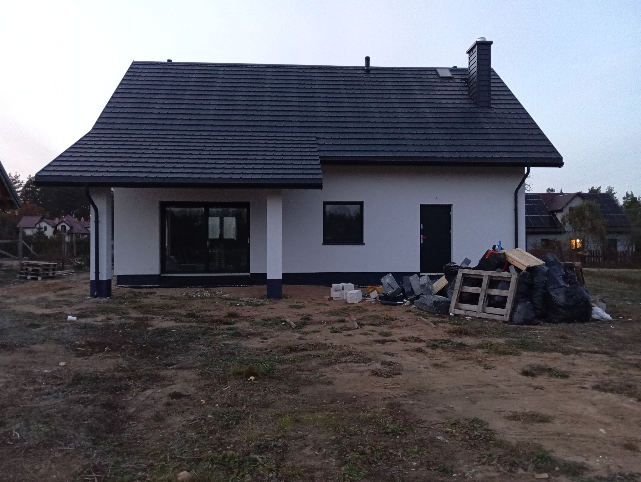 Biały dom jednorodzinny z ciemną dachówką, widok z zewnątrz, teren wokół domu w trakcie zagospodarowania, widoczne materiały budowlane i odpady.