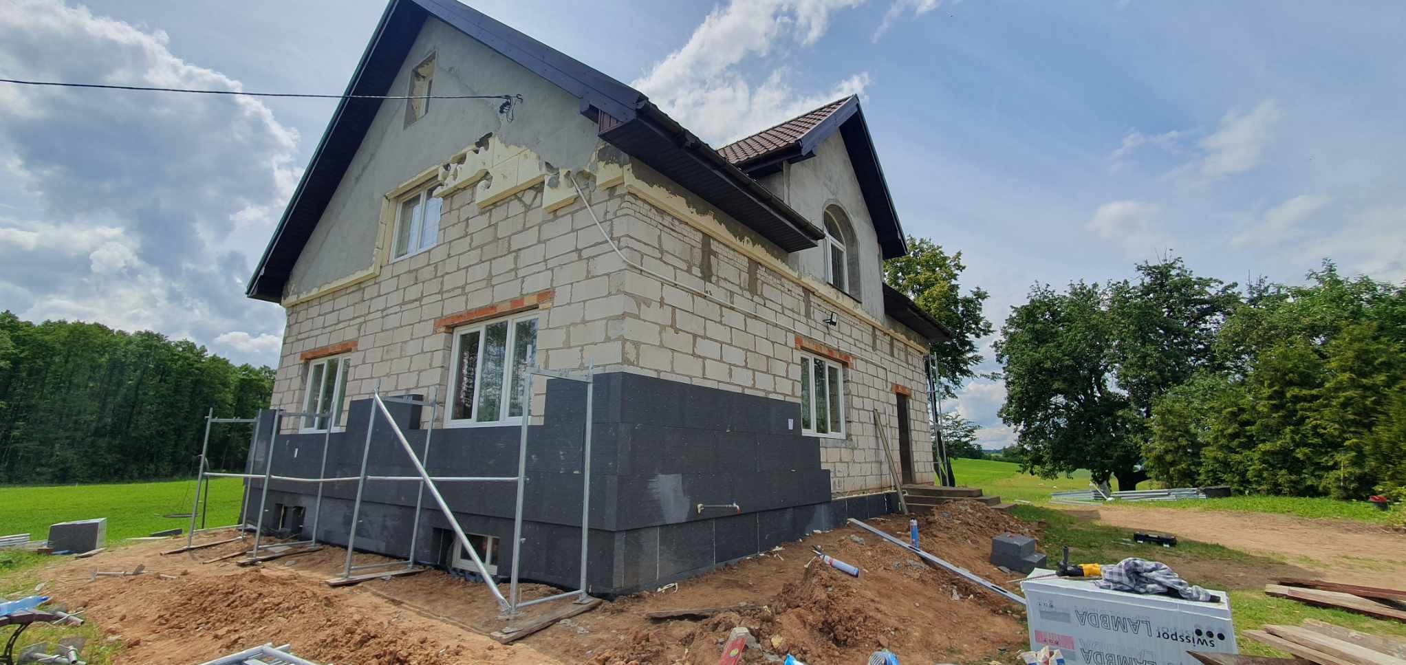 Ocieplenie grafitowym styropianem fragmentu elewacji domu jednorodzinnego z białego bloczka. Widoczne rusztowania, narzędzia i materiały budowlane.