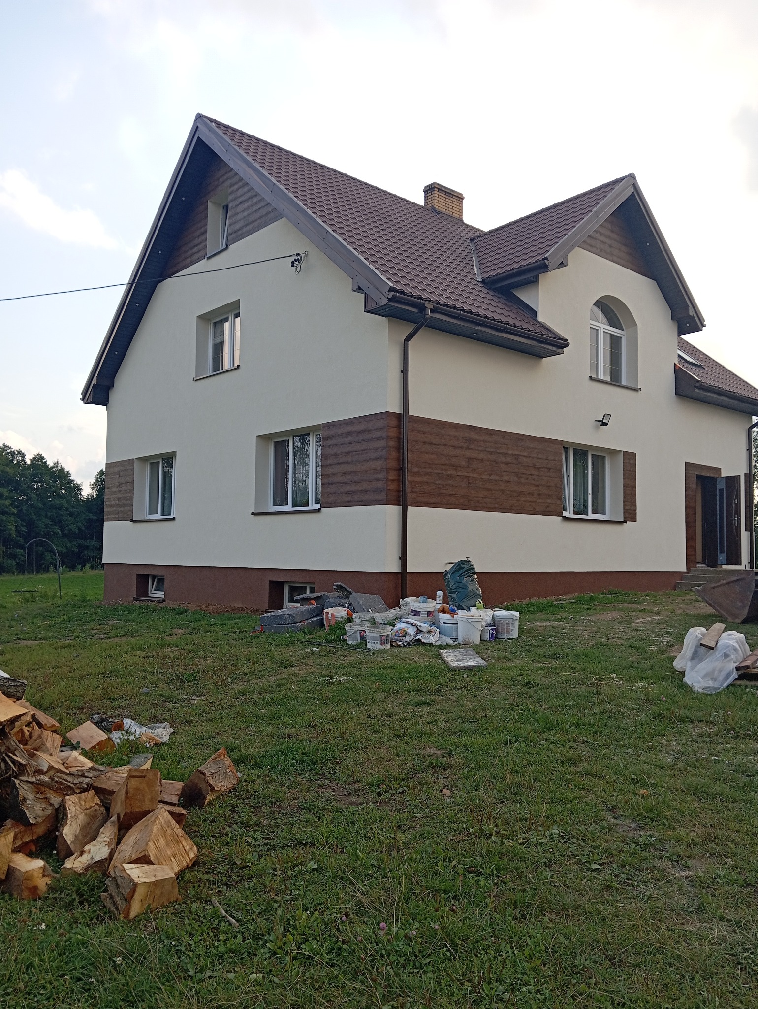 Dom jednorodzinny po renowacji elewacji, z elementami dekoracyjnymi imitującymi drewno. Widoczne puszki po farbach, świeżo pomalowany budynek z brązowym dachem.