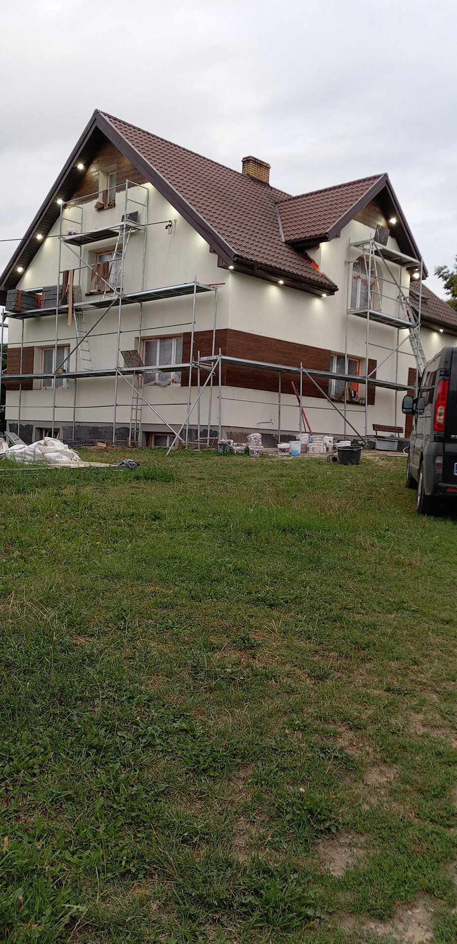 Dom z rusztowaniami podczas prac elewacyjnych. Część ściany z brązowymi panelami, reszta w jasnym kolorze. Widoczne worki i narzędzia.