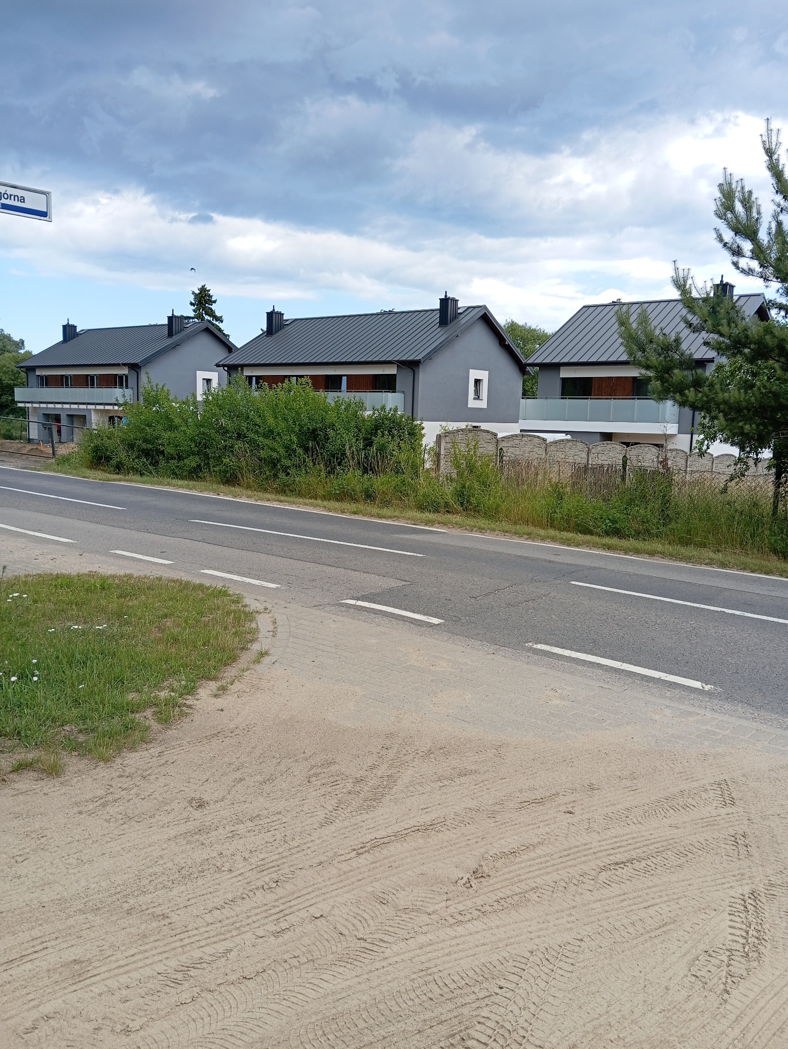 Trzy identyczne, szare domy jednorodzinne z ciemnymi dachami, oddzielone niskim, kamiennym ogrodzeniem, widziane z perspektywy ulicy w pochmurny dzień.