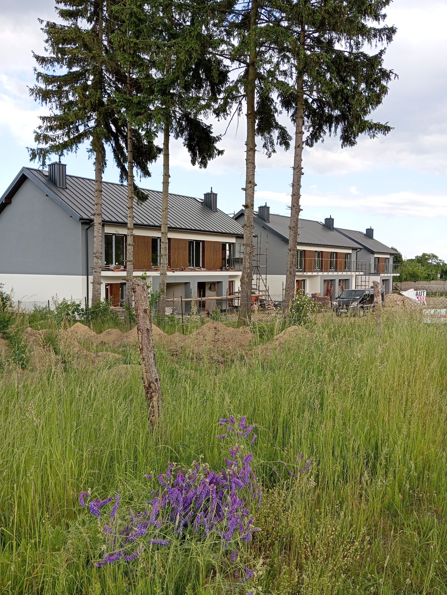 Nowoczesne domy w zabudowie szeregowej z szarą elewacją i drewnianymi elementami, otoczone zielenią i drzewami. Częściowo widoczne rusztowania.