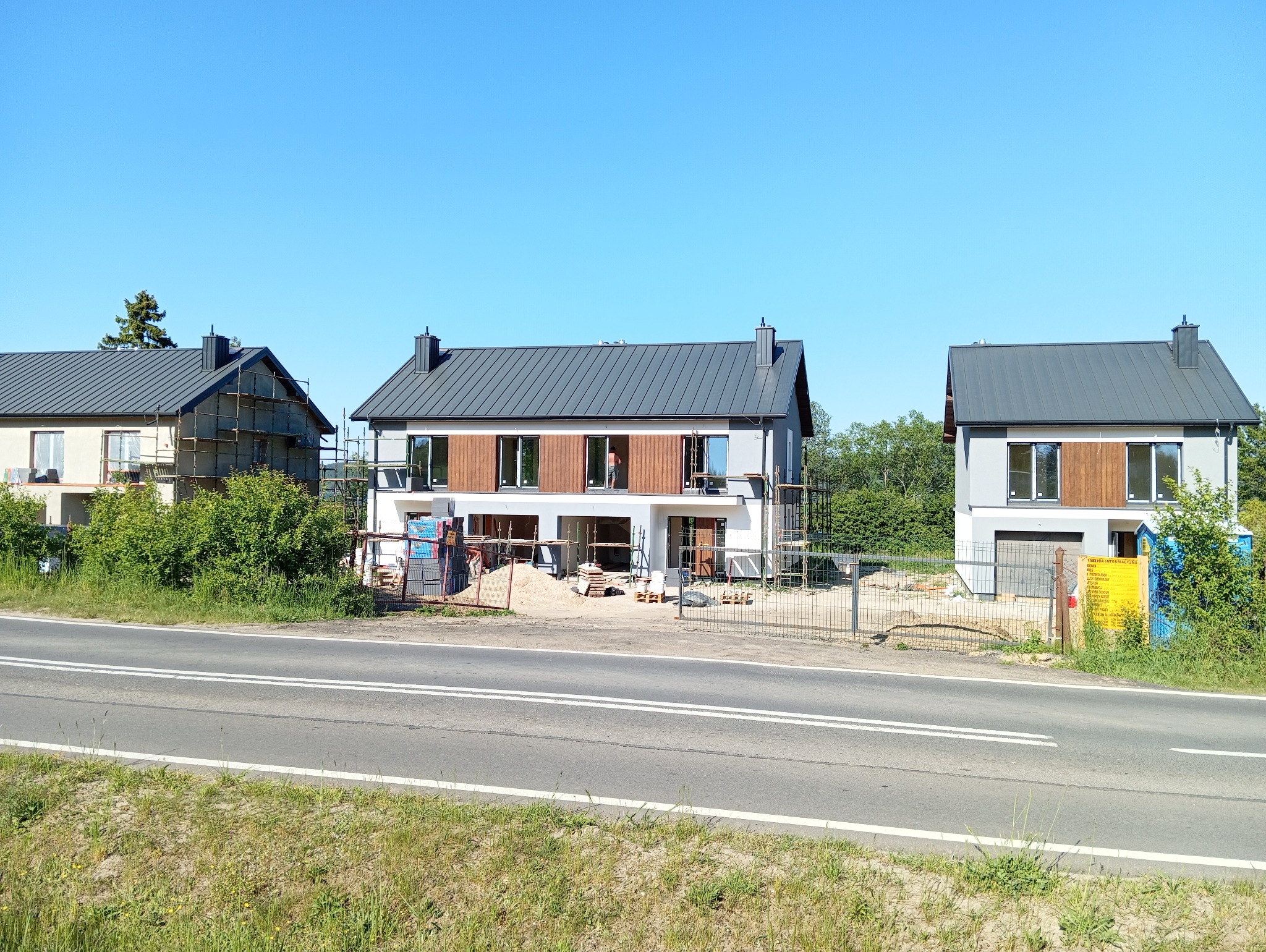 Trzy domy w różnym stadium prac elewacyjnych. Jeden dom w rusztowaniach, drugi z częściowo ukończoną elewacją, trzeci gotowy. Widok zza drogi.