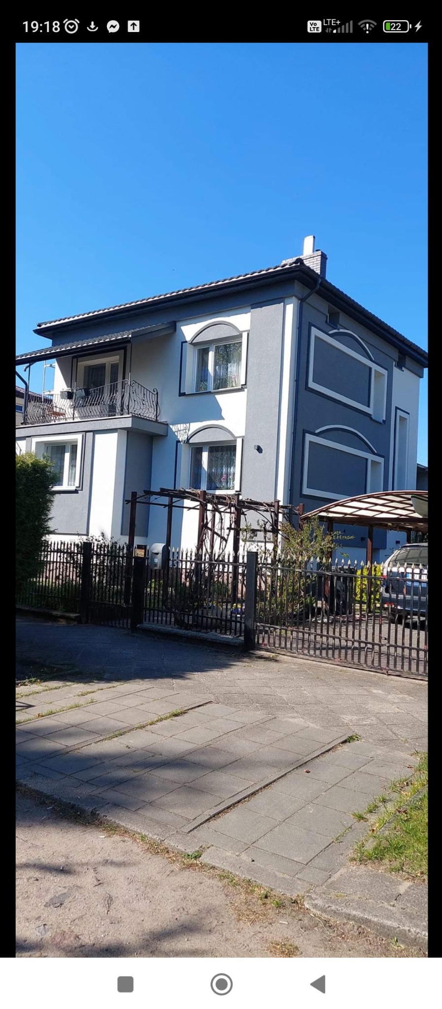 Dwukondygnacyjny dom z szaro-białą elewacją, balkonem z kutą balustradą i dekoracyjnymi obramowaniami okien. Widok z zewnątrz, brama i podjazd.