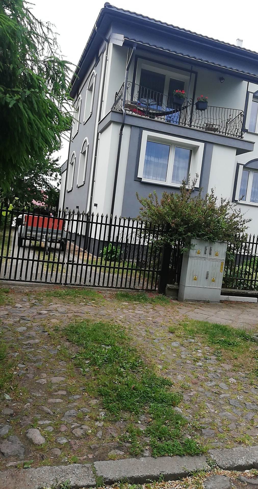 Dwukondygnacyjny dom po renowacji elewacji w odcieniach szarości i bieli, z balkonem z kutą balustradą, widoczny zza czarnego ogrodzenia.