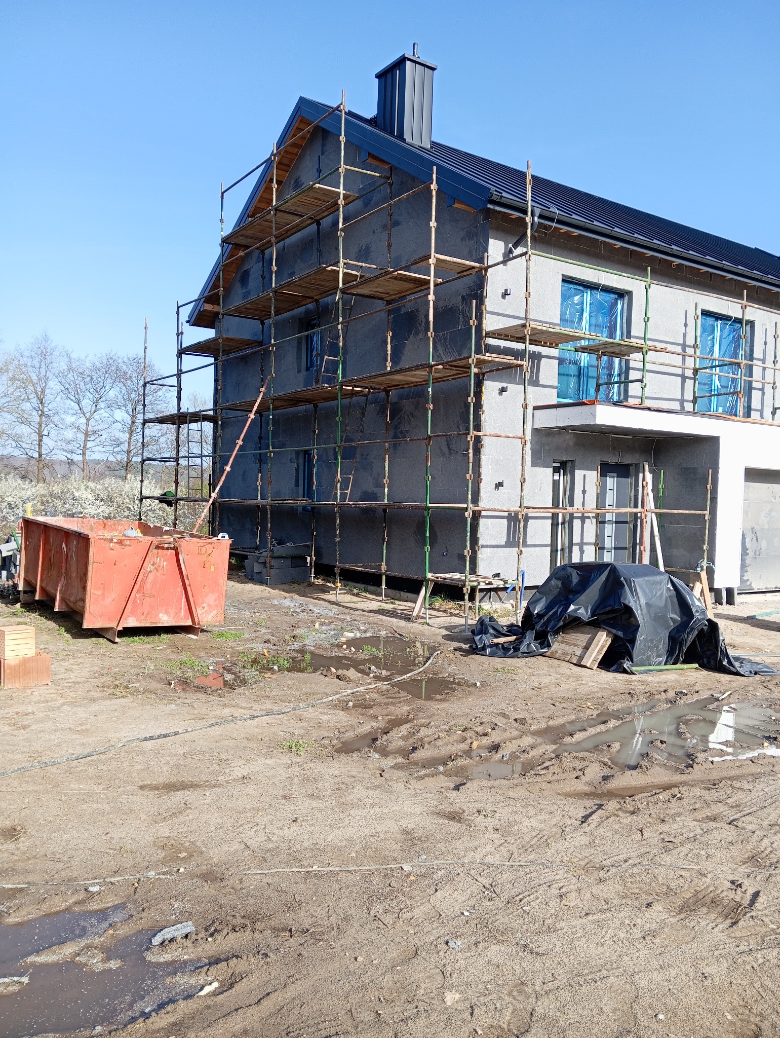 Dom w trakcie ocieplania elewacji, otoczony rusztowaniami. Widoczny kontener na odpady i materiały budowlane. Okna zabezpieczone folią. Teren wokół placu budowy.