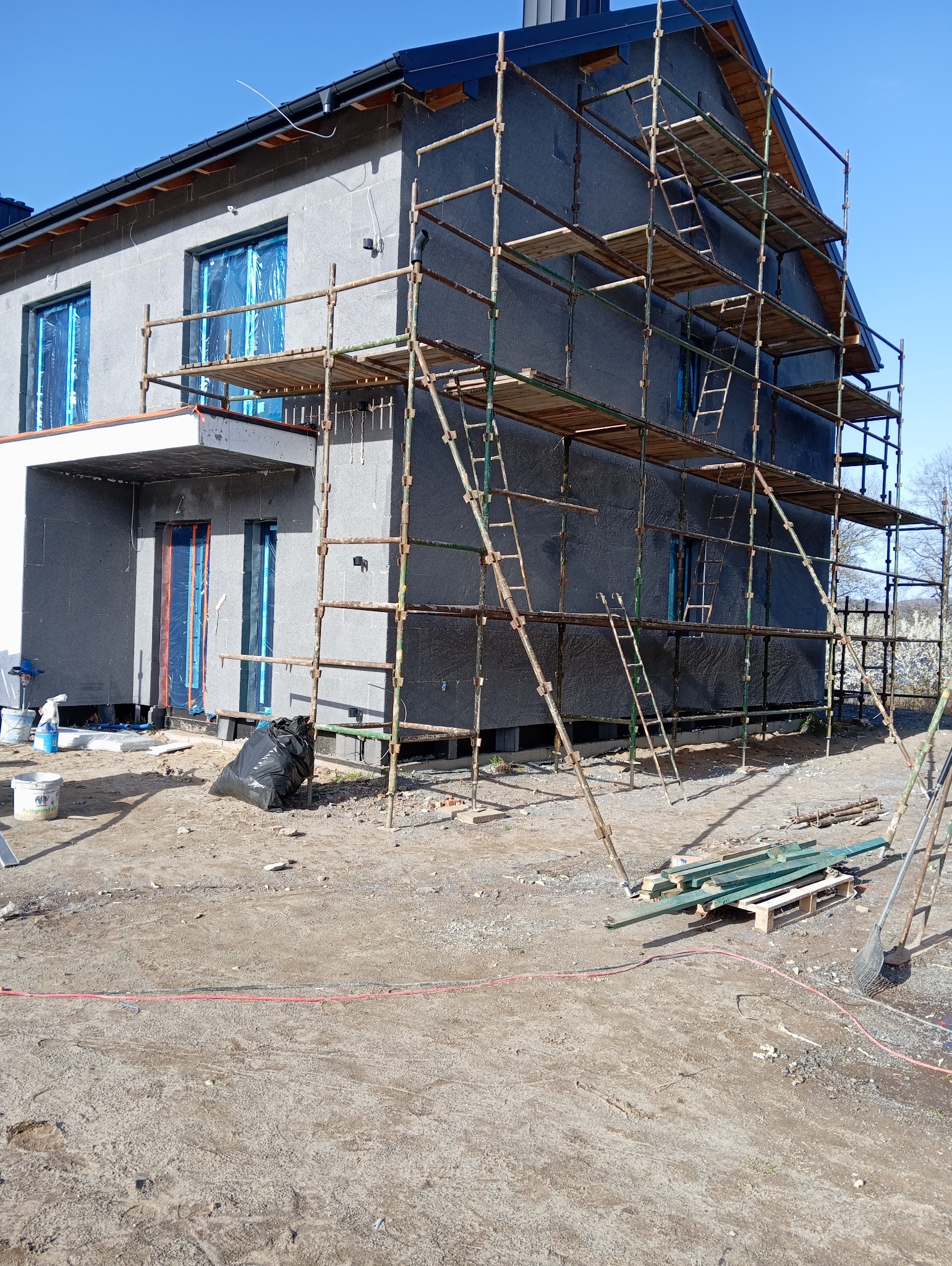 Dom w trakcie ocieplania elewacji. Widoczne rusztowania, materiały izolacyjne i okna zabezpieczone folią. Na ziemi narzędzia i odpady budowlane.