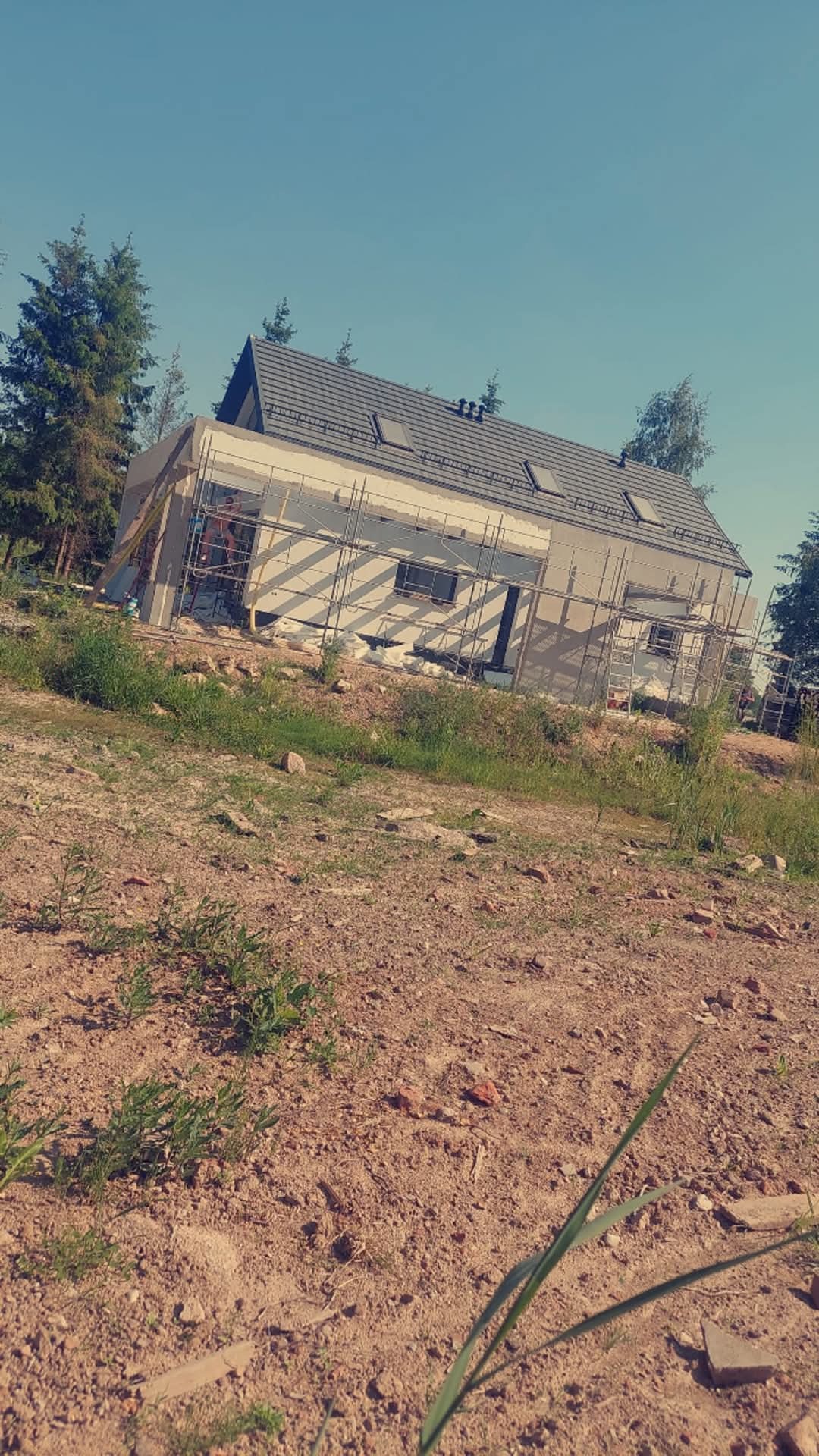 Dom jednorodzinny w trakcie ocieplania, widoczne rusztowania. Dach pokryty szarą dachówką. Tło stanowi błękitne niebo i zieleń drzew.