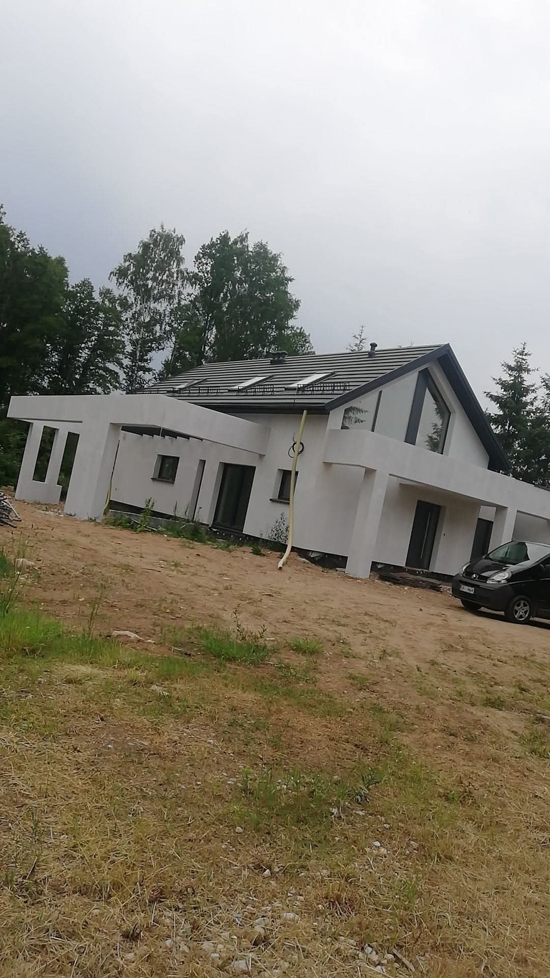 Nowoczesny dom jednorodzinny z białą elewacją, czarną dachówką i dużymi oknami, widziany z poziomu gruntu na tle drzew i pochmurnego nieba.