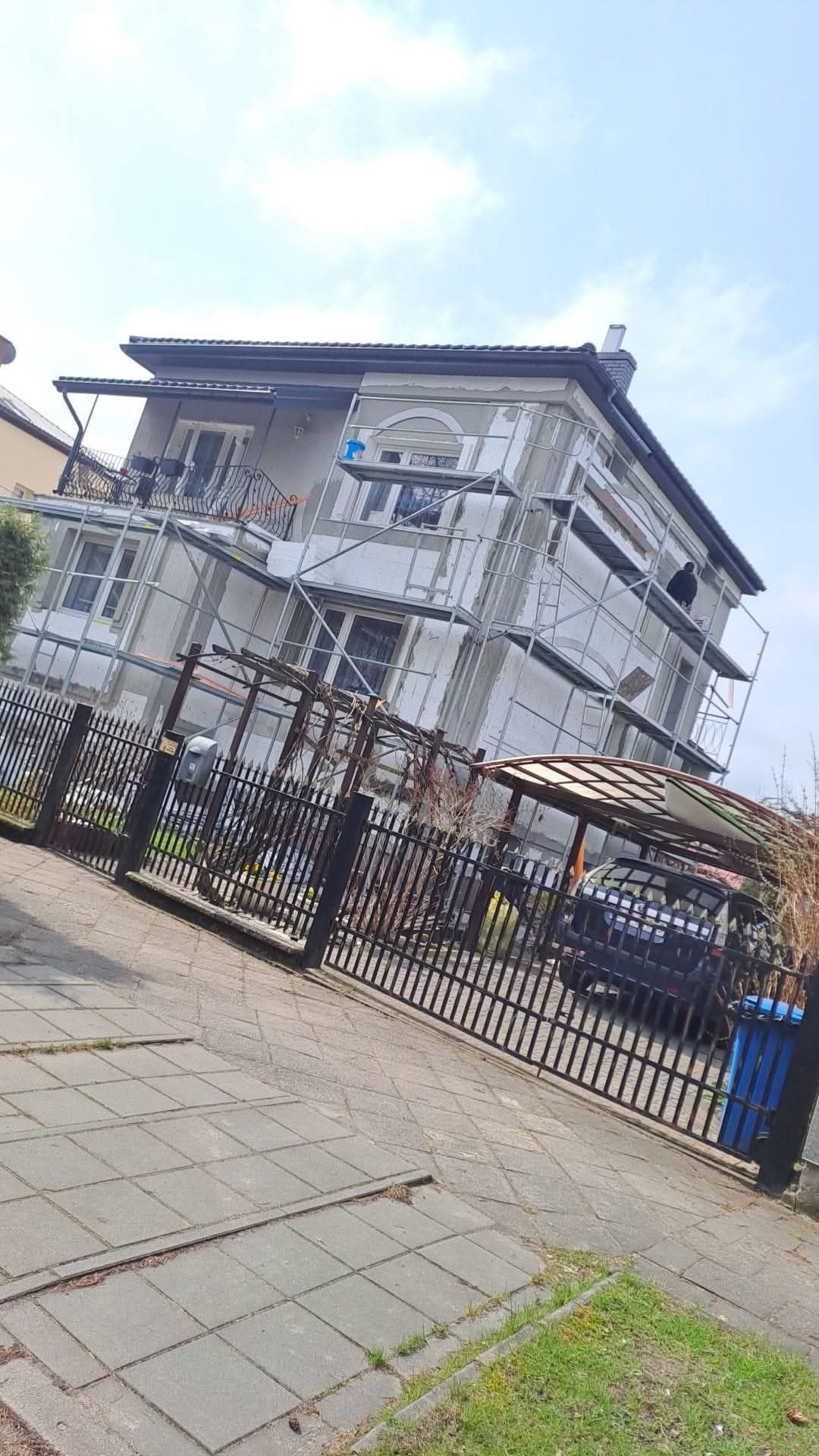 Dom w trakcie renowacji elewacji, z rusztowaniami i pracownikiem na górze. Widoczne ogrodzenie, chodnik i samochód w tle.