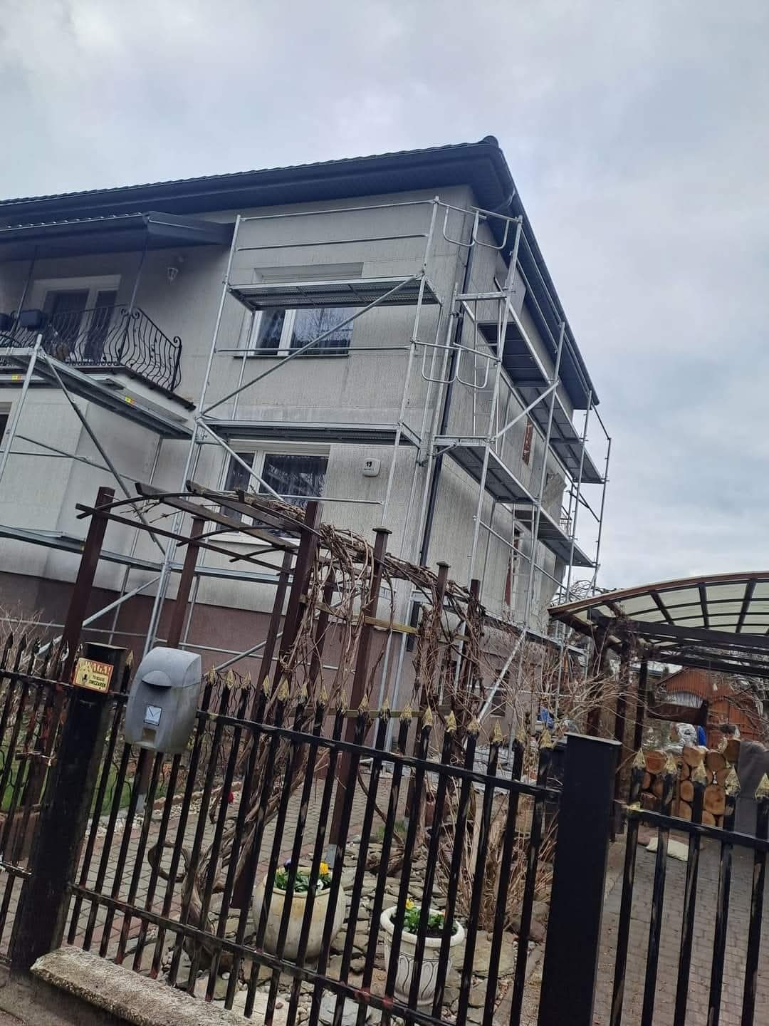 Dom w trakcie ocieplania, widoczne rusztowania i szara elewacja. Na pierwszym planie ozdobny płot z roślinnością i skrzynka pocztowa.