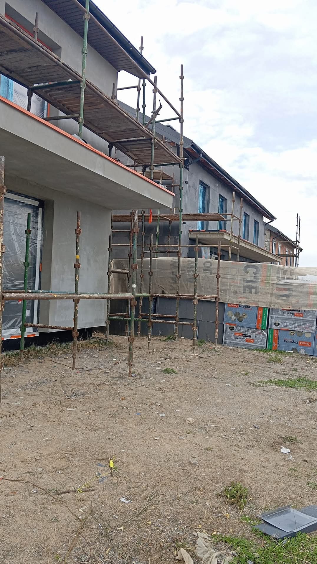 Dom w trakcie ocieplania: szare ściany, rusztowania, styropian. Częściowo otynkowana elewacja z oknami zabezpieczonymi folią. Widoczne materiały izolacyjne.