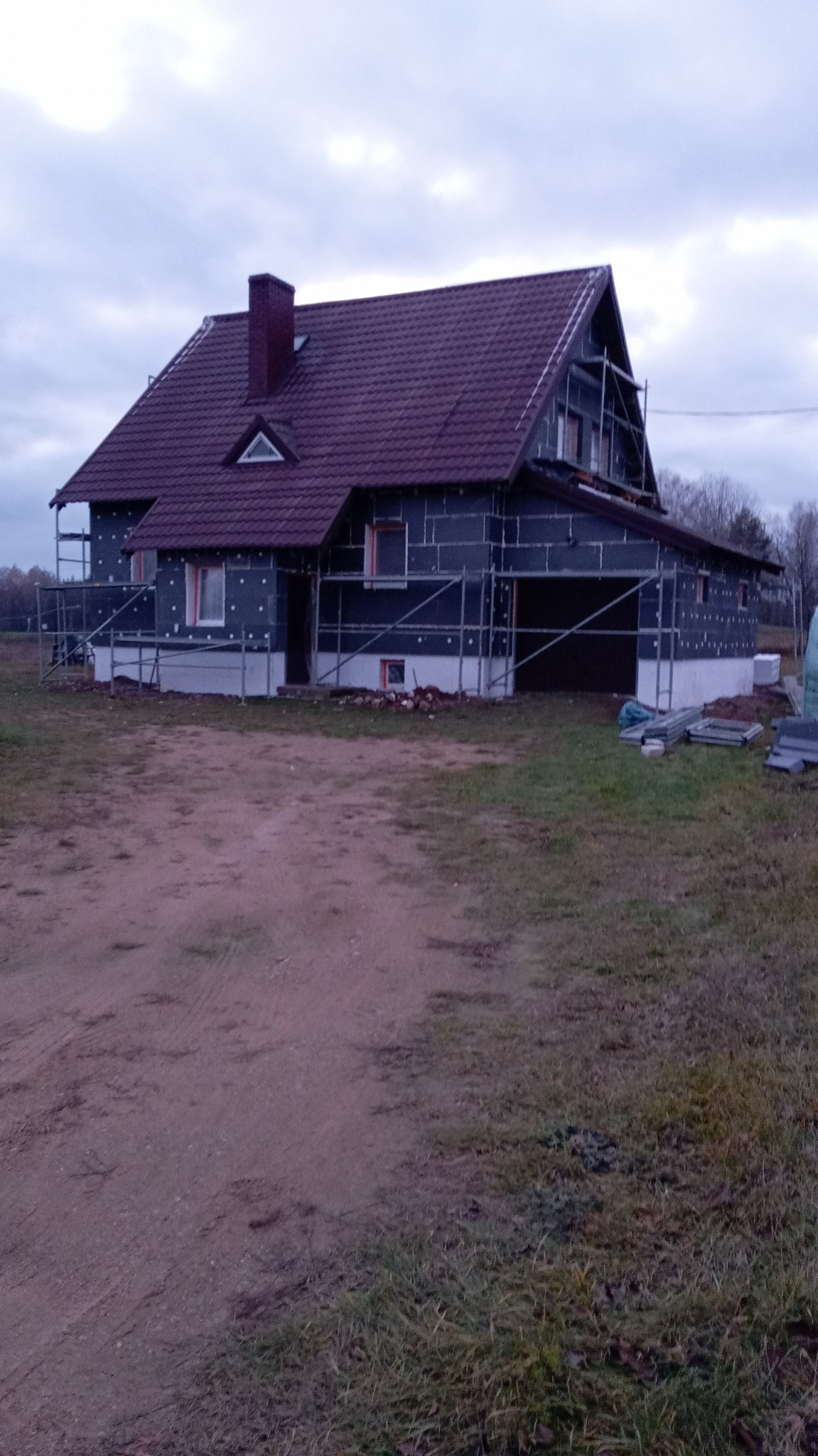 Dom jednorodzinny w trakcie ocieplania, widoczne rusztowania i płyty izolacyjne na elewacji. Dach pokryty bordową dachówką. Pochmurne niebo.