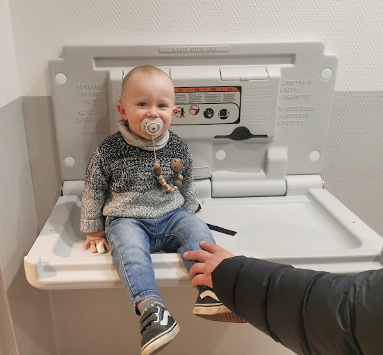 Uśmiechnięty chłopiec siedzi na rozkładanym przewijaku w publicznej toalecie, z drewnianym gryzakiem na szyi i smoczkiem w ustach, wspierany przez dłoń osoby dorosłej.