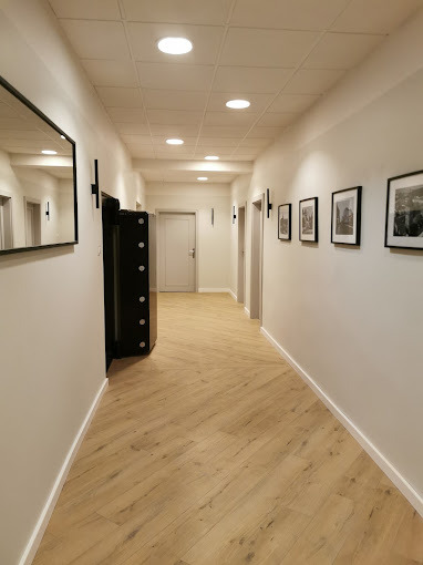 Korytarz biurowy z drewnianą podłogą ułożoną we wzór jodełki, czarnym sejfem z boku, oświetleniem punktowym i obrazami na ścianie.