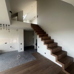 RENOVA CONSTRUCTION SPÓŁKA Z OGRANICZONĄ ODPOWIEDZIALNOŚCIĄ - Wnętrze w trakcie remontu: widoczne surowe ściany, częściowo położona podłoga z szarego lastryko i drewnianych paneli, oraz drewniane schody bez poręczy.