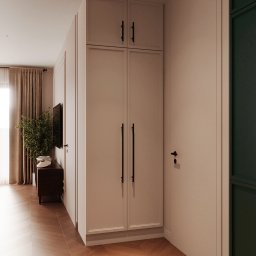 RENOVA CONSTRUCTION SPÓŁKA Z OGRANICZONĄ ODPOWIEDZIALNOŚCIĄ - Wnętrze mieszkania z zabudową szafy w jasnym odcieniu, ciemnymi uchwytami i parkietem w jodełkę; w tle widoczny telewizor, roślina doniczkowa i zasłonięte okno.