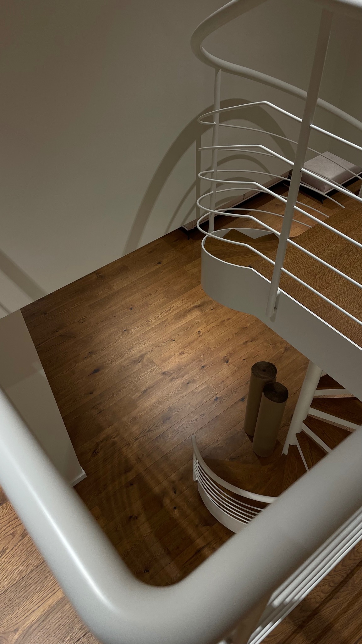 Widok z góry na odnowioną klatkę schodową z drewnianymi schodami i białą, spiralną balustradą. Na podłodze leżą dwie rolki papieru lub folii zabezpieczającej.