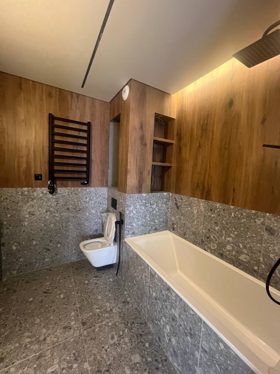 Nowoczesna łazienka z drewnianymi panelami, szarą mozaiką i białymi sanitariatami. Grzejnik drabinkowy w kolorze czarnym, minimalistyczne oświetlenie sufitowe.