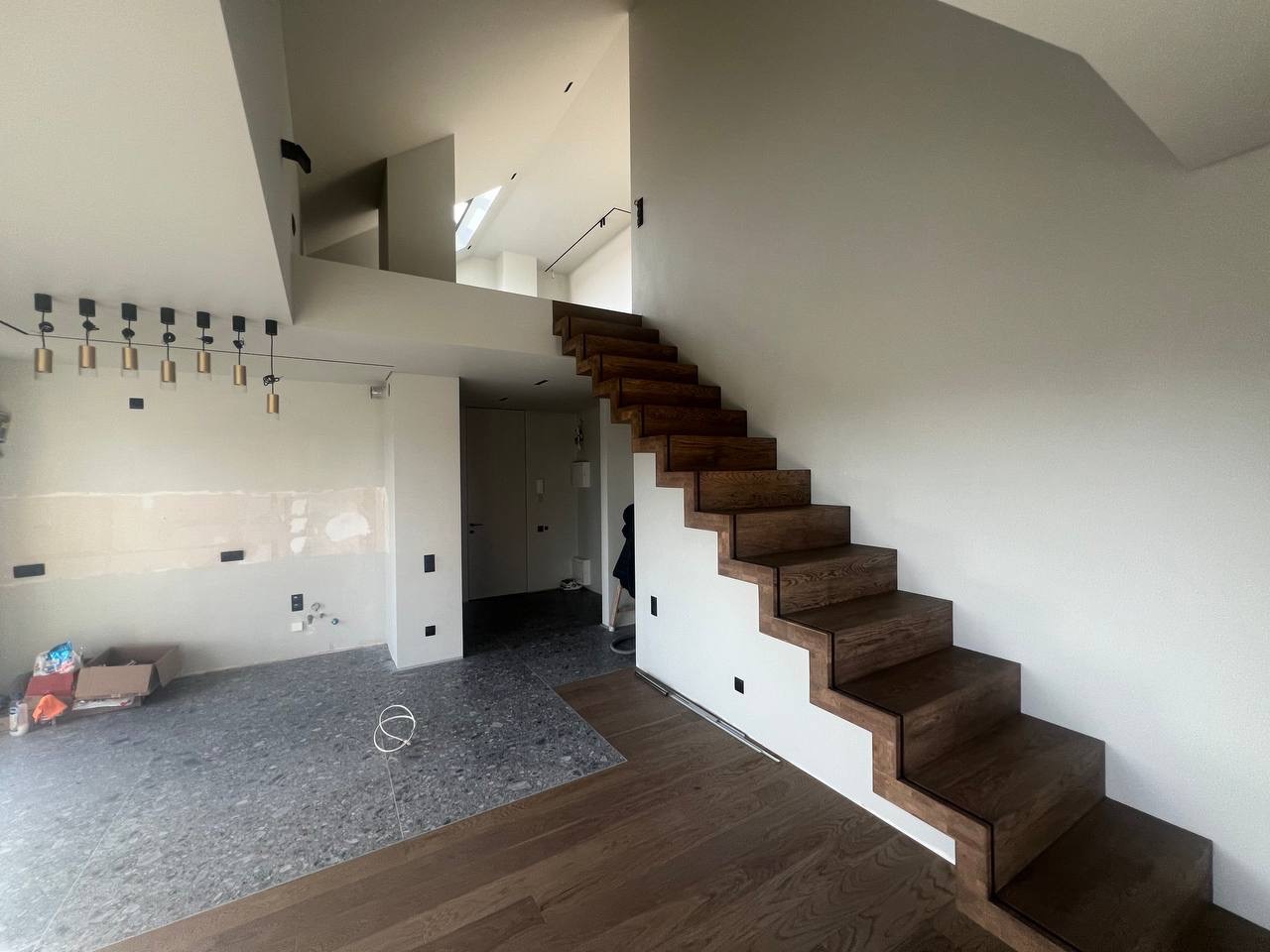 Wnętrze w trakcie remontu: widoczne surowe ściany, częściowo położona podłoga z szarego lastryko i drewnianych paneli, oraz drewniane schody bez poręczy.