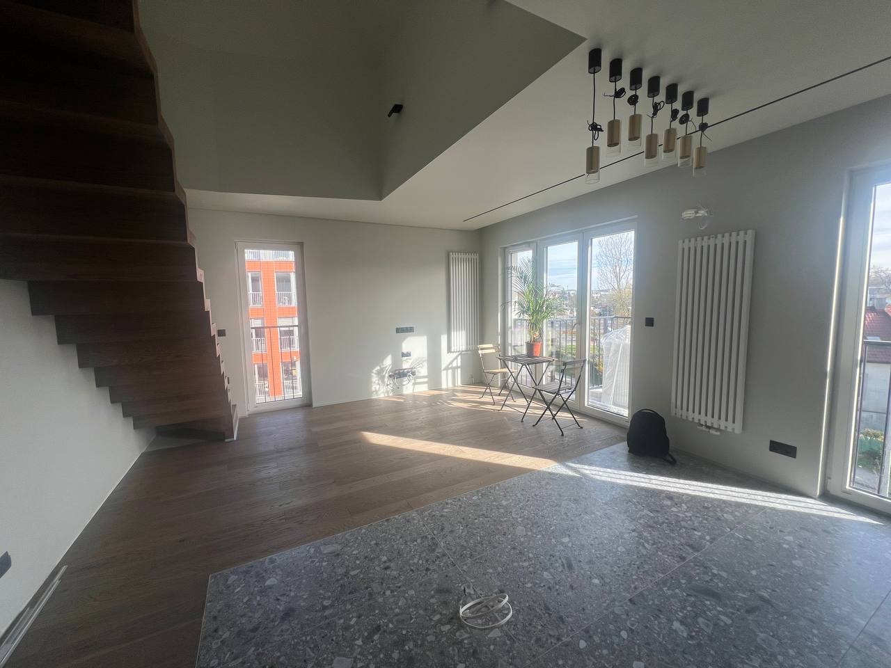 Jasne, przestronne wnętrze z drewnianymi schodami, parkietem i szarą podłogą z lastryko, widoczne grzejniki, okna balkonowe i nowoczesne oświetlenie sufitowe.