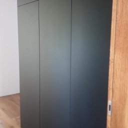 Tomasz Hawro - Czarna, minimalistyczna zabudowa szafy z ukrytymi uchwytami we Wrocławiu, dopasowana do drewnianej podłogi i framugi drzwi, z widocznymi subtelnymi liniami podziału frontów.