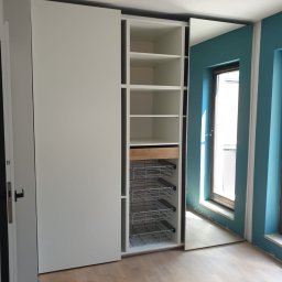 Tomasz Hawro - Biała szafa z przesuwnymi drzwiami, widoczne półki, wysuwana szuflada z jasnego drewna i metalowe kosze, na podłodze panele, ściana pomalowana na turkusowo.