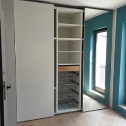 Biała szafa z przesuwnymi drzwiami, widoczne p&oacute;łki, wysuwana szuflada z jasnego drewna i metalowe kosze, na podłodze panele, ściana pomalowana na turkusowo.