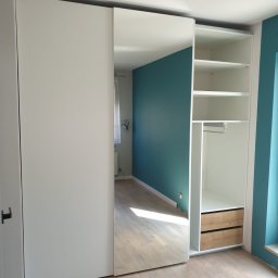 Tomasz Hawro - Biała szafa z przesuwnymi drzwiami z lustrem, widoczne półki i dwie szuflady z jasnego drewna, na podłodze panele w odcieniach beżu, ściana w kolorze turkusowym.