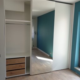 Tomasz Hawro - Biała szafa przesuwna z lustrem i drewnianymi szufladami, widok na wnętrze szafy i odbicie pokoju z turkusową ścianą.