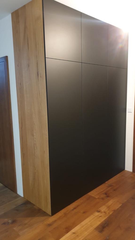 Czarna szafa z drewnianym bokiem, widoczna na tle drewnianej podłogi i białej ściany, z delikatnym odbiciem światła na frontach.