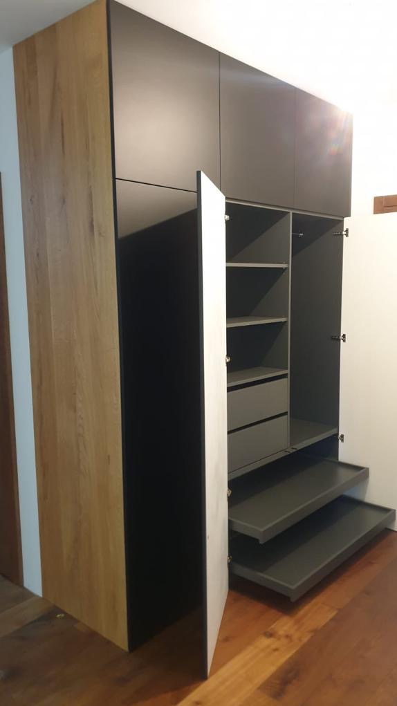 Wysoka szafa z frontami w kolorze grafitowym i elementami z drewna w odcieniu dębu, z otwartymi drzwiami ukazującymi szare półki i szuflady oraz wysuwane platformy.