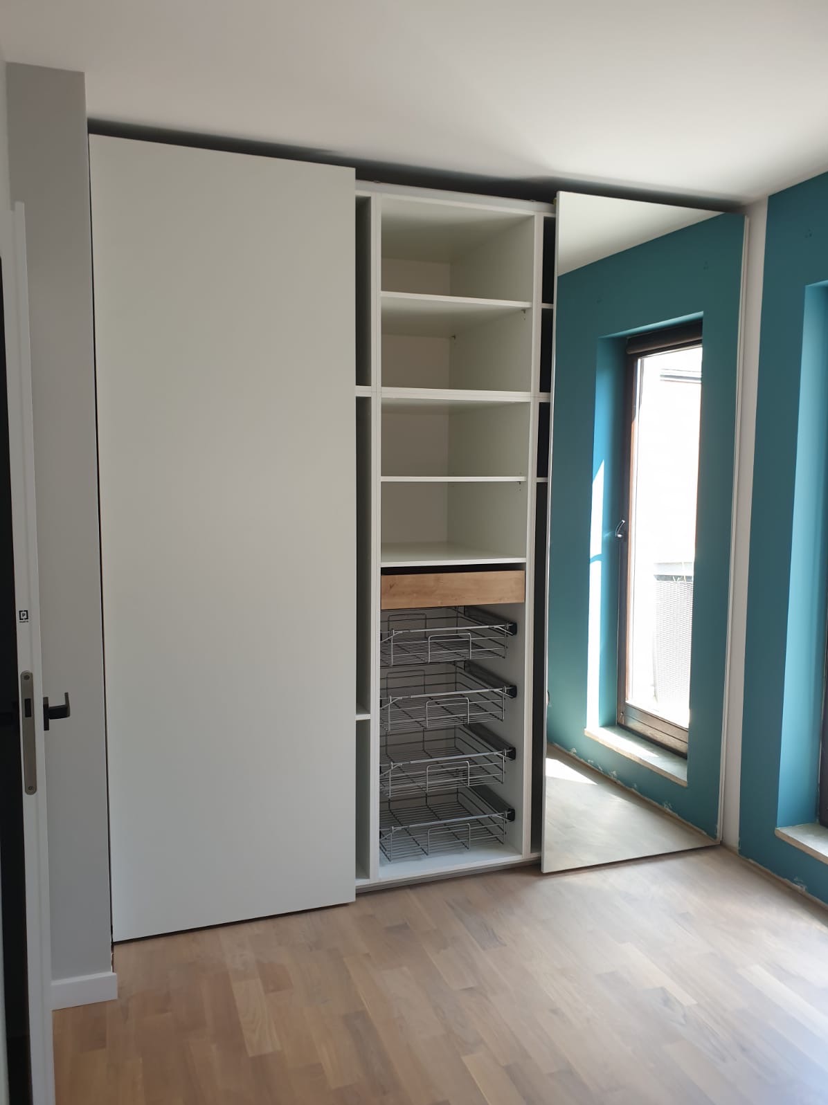 Biała szafa z przesuwnymi drzwiami, widoczne półki, wysuwana szuflada z jasnego drewna i metalowe kosze, na podłodze panele, ściana pomalowana na turkusowo.